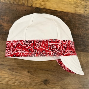 Puede incluir: Una gorra de soldador blanca con una banda ancha de tela roja con un patrón de cachemira en blanco y negro. La gorra tiene una corona redondeada construida con varios paneles y una visera corta y rígida que muestra un forro de cachemira rojo.