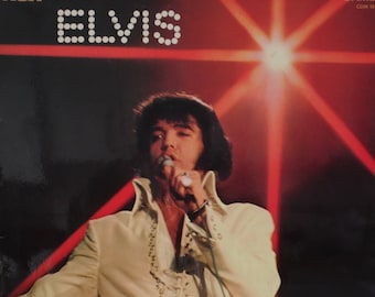 Può includere: Una copertina di un disco in vinile vintage con Elvis Presley in un abito bianco, che canta in un microfono. Lo sfondo è un palcoscenico rosso e nero con i riflettori. La copertina riporta la scritta "Elvis" in grandi lettere bianche e "Camden" in lettere bianche più piccole.