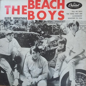Può includere: Foto in bianco e nero dei membri della band The Beach Boys in piedi intorno a un'auto. La copertina dell'album presenta il nome della band in lettere rosse e il titolo "Good Vibrations" in lettere nere. Il logo della Capitol Records è nell'angolo in alto a destra. Il testo "Tell me why the times they are a-changin' devoted to you" è stampato nell'angolo in alto a destra.