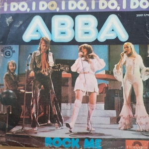 Pode incluir: Uma capa de disco de vinil vintage com a banda ABBA. A capa é roxa e branca com uma foto dos membros da banda. O título do disco é "I Do, I Do, I Do, I Do, I Do" e o título da música é "Rock Me".
