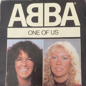 Pode incluir: Capa do álbum de vinil vintage "One of Us" do ABBA. A capa do álbum apresenta o nome da banda em letras brancas grandes, com o título da música abaixo. Retratos de dois membros da banda são exibidos abaixo.