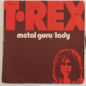 Pode incluir: Um disco de vinil vermelho e branco com o texto "T.Rex" em letras vermelhas grandes. O disco é intitulado "Metal Guru/Lady" e apresenta uma foto em preto e branco de Marc Boland na capa.