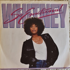May include: A vinyl record cover featuring Whitney Houston with the text WHITNEY in large purple letters and So Emotional in red script. The artist wears a black leather jacket over a white tank top and blue jeans, with a cross necklace.