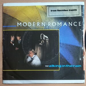 Könnte beinhalten: Albumcover für "Walking in the Rain" von Modern Romance. Das Cover zeigt ein Foto der Bandmitglieder in dunklen Anzügen und einem weißen Hemd. Der Hintergrund ist eine Mischung aus Blau, Gelb und Schwarz. Der Text enthält "free flexidisc inside."