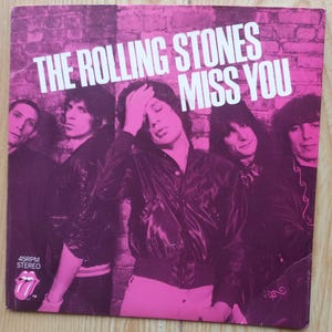 Pode incluir: Capa do álbum do single "Miss You" dos Rolling Stones. A imagem apresenta a banda posando em frente a uma parede de tijolos rosa e roxa. O logotipo da língua dos Rolling Stones também é visível.