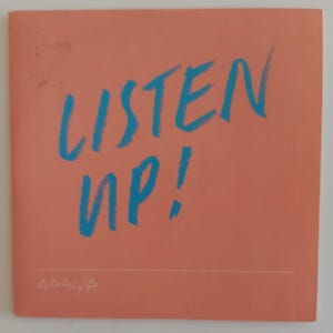 May include: A square, coral-coloured card with the words "LISTEN UP!" in bright blue, handwritten-style lettering. A thin white line runs horizontally across the bottom, with small, illegible text below it.