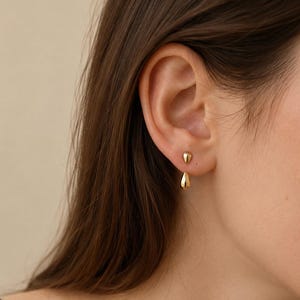 May include: A gold earring featuring a small, smooth, teardrop-shaped stud at the earlobe and a matching, slightly larger teardrop-shaped charm suspended directly below it, creating a layered, minimalist aesthetic with a polished, reflective finish.