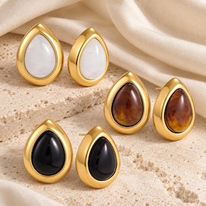 May include: Three pairs of teardrop-shaped stud earrings featuring gold-toned metal frames. One pair contains white marbled inserts, one pair contains brown tortoiseshell-patterned inserts, and one pair contains solid black smooth inserts.