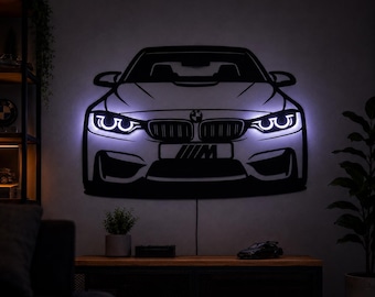 May include: A black metal wall art piece shaped like the front of a BMW M car, featuring integrated white LED backlighting that illuminates the headlights and the car's outline. The design includes the BMW logo and an M badge on the front bumper.