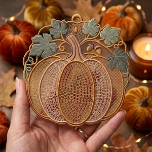 May include: A circular pumpkin-shaped ornament featuring intricate golden-brown lace embroidery with a mesh pattern. The design includes sage green leaves, small brown acorns, and a delicate spiderweb detail at the top, finished with a small hanging loop.