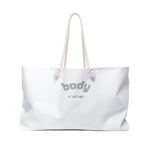 May include: A white tote bag with two long rope handles and the text body n'align printed in black on the front. The bag features a rectangular shape with a clean, smooth surface and knotted rope attachments at the top of the main compartment.