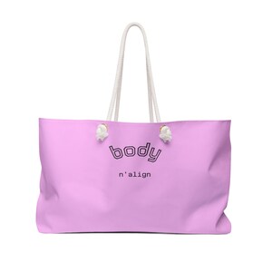 May include: A light pink tote bag with white rope handles and gold-toned hardware. The front features the word "body" in a black outlined varsity-style font above the text "n'align" in a smaller, simple black sans-serif typeface.