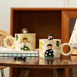 Peut inclure: Deux tasses en céramique de couleur crème avec de grandes anses rondes et des couvercles assortis. Chaque couvercle comporte une petite figurine 3D. Les tasses sont décorées d'illustrations peintes de personnages en vêtements à pois.