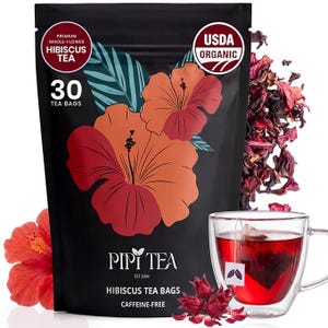 May include: A black pouch of Pipi Tea hibiscus tea bags featuring large orange and red hibiscus flower illustrations, a USDA Organic seal, and text stating 30 tea bags. A glass mug filled with red hibiscus tea sits next to loose dried petals.