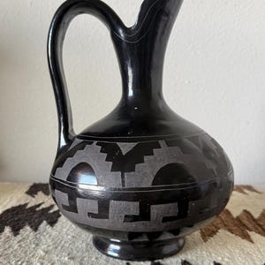 May include: A black ceramic pitcher with a glossy finish and a matte gray geometric pattern encircling the rounded body. The vessel features a curved handle, a narrow neck, and a flared spout, showcasing a contrast between the shiny and matte surfaces.