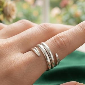 Puede incluir: Un anillo de plata con diseño de bypass que presenta dos bandas lisas y redondeadas que se envuelven alrededor del dedo. El metal tiene un acabado pulido y reflectante, y los extremos de las bandas terminan en puntas redondeadas.