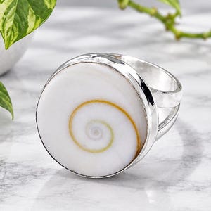 Könnte beinhalten: Ein Silberring mit einer runden weißen Muscheleinlage, die ein natürliches goldenes Spiralmuster aufweist. Der Ring hat eine polierte Metallschiene und wird zusammen mit dem Text Laxmijewellersco und einem Lotusblüten-Symbol gezeigt.