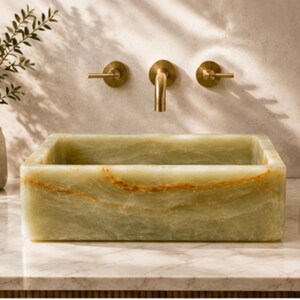 May include: A rectangular vessel sink crafted from light green onyx stone with prominent orange veining throughout. The sink features a smooth, polished finish and is paired with a wall-mounted, brushed brass faucet set consisting of a central spout and two handles.