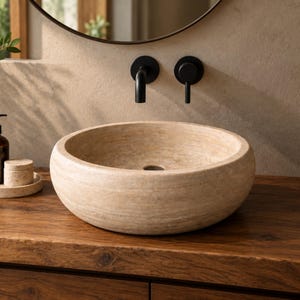 Puede incluir: Un lavabo de piedra natural beige, de forma redonda, con una superficie texturizada y porosa y un orificio de drenaje central. El lavabo se asienta sobre una encimera de madera oscura, junto a un grifo de pared negro mate y un mando negro.