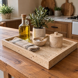 May include: A rectangular beige travertine tray holding a glass oil bottle with a cork stopper, a small potted plant, a matching stone bowl with a gold spoon, a lidded stone jar, and a folded beige linen napkin with dark stripes.