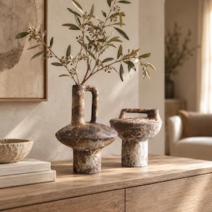 May include: Two textured ceramic vases with a weathered brown and cream finish. The taller vase features a central handle and a wide, rounded body, while the shorter vase has a flat top handle and a cylindrical base, holding green leafy branches.