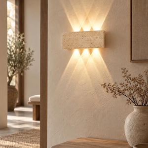 Peut inclure: Une applique murale rectangulaire fabriquée en pierre de travertin beige poreuse. Le luminaire est fixé au mur et émet une lumière chaude et vive en trois faisceaux distincts vers le haut et trois vers le bas, créant un effet d'étoile.