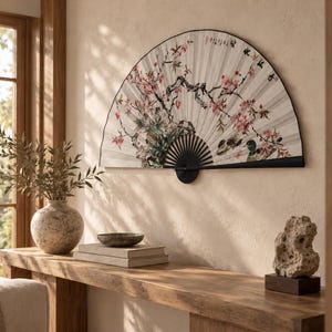Pode incluir: Um grande leque dobrável semicircular montado na parede, com fundo branco e uma ilustração detalhada de flores de cerejeira cor-de-rosa, ramos escuros e caligrafia preta. O leque tem uma estrutura preta e um cabo preto na base.