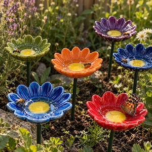 May include: A collection of six ceramic flower-shaped garden stakes in green, orange, purple, dark blue, and red. Each flower features a yellow centre and serves as a basin for water, with bees and a butterfly resting on the petals.