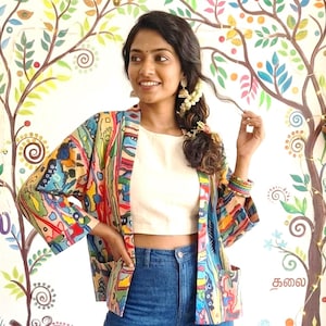 Peut inclure: Veste kimono ouverte aux motifs colorés, avec des motifs abstraits dans les tons de bleu, rouge, jaune et vert. Portée sur un crop top blanc et un jean bleu. Le fond présente une fresque murale.