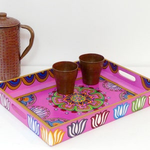 Peut inclure: Un plateau décoratif en bois rose avec un motif floral et deux petites tasses en cuivre. Le plateau a une bordure dorée et des motifs de tulipes. Une cruche en cuivre à la surface texturée est également présente.