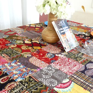 Peut inclure: Une nappe de patchwork color&eacute;e avec une vari&eacute;t&eacute; de motifs et de couleurs, notamment le rouge, le vert, le bleu et le jaune. Un vase avec des fleurs blanches et un livre intitul&eacute; "Les Fils de la Krishna" sont sur la table.