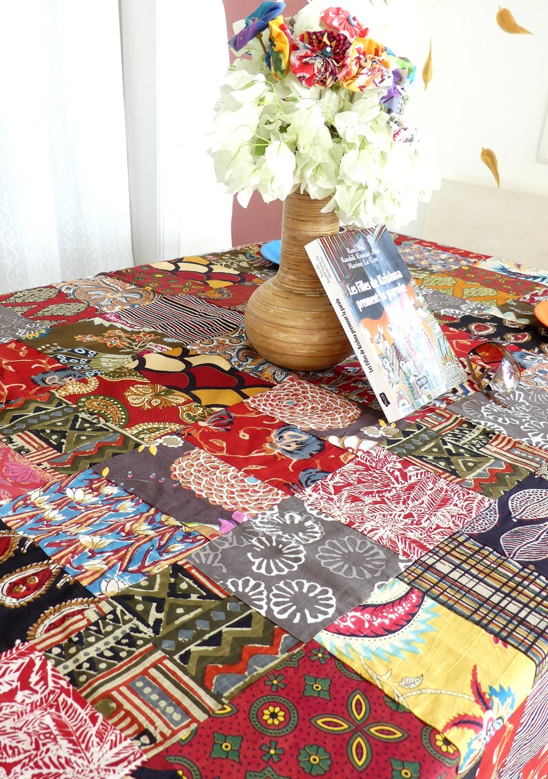 Peut inclure: Une nappe de patchwork color&eacute;e avec une vari&eacute;t&eacute; de motifs et de couleurs, notamment le rouge, le vert, le bleu et le jaune. Un vase avec des fleurs blanches et un livre intitul&eacute; "Les Fils de la Krishna" sont sur la table.