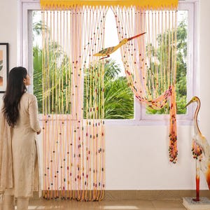 Könnte beinhalten: Ein Fenstervorhang aus gelben und orangefarbenen Schnüren mit bunten Perlen. Ein dekorativer Vogel ist am Vorhang befestigt. Eine dekorative Vogelstatue steht rechts vom Fenster. Eine Frau steht vor dem Fenster.