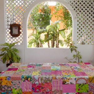 K&ouml;nnte beinhalten: Eine bunte Patchwork-Tischdecke mit verschiedenen Mustern und Farben, darunter Pink, Gelb, Gr&uuml;n und Blau. Die Tischdecke liegt auf einem Tisch in einem Raum mit einem wei&szlig;en Gitterfenster und Blick auf &uuml;ppiges Gr&uuml;n.