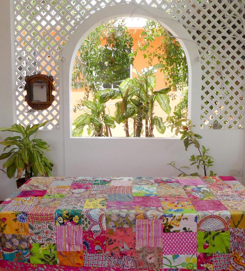 K&ouml;nnte beinhalten: Eine bunte Patchwork-Tischdecke mit verschiedenen Mustern und Farben, darunter Pink, Gelb, Gr&uuml;n und Blau. Die Tischdecke liegt auf einem Tisch in einem Raum mit einem wei&szlig;en Gitterfenster und Blick auf &uuml;ppiges Gr&uuml;n.