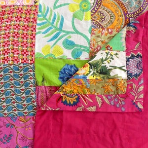 K&ouml;nnte beinhalten: Eine Patchwork-Decke mit rosa R&uuml;ckseite und einem bunten Patchwork-Design mit Blumenmustern, geometrischen Formen und Paisley-Drucken.