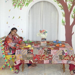 Peut inclure: Une femme est assise à une table recouverte d'une nappe en patchwork colorée. La table est dressée avec quatre assiettes et tasses colorées. La femme porte une chemise à motifs rouges et bleus. L'arrière-plan présente une peinture murale d'un arbre avec des oiseaux.