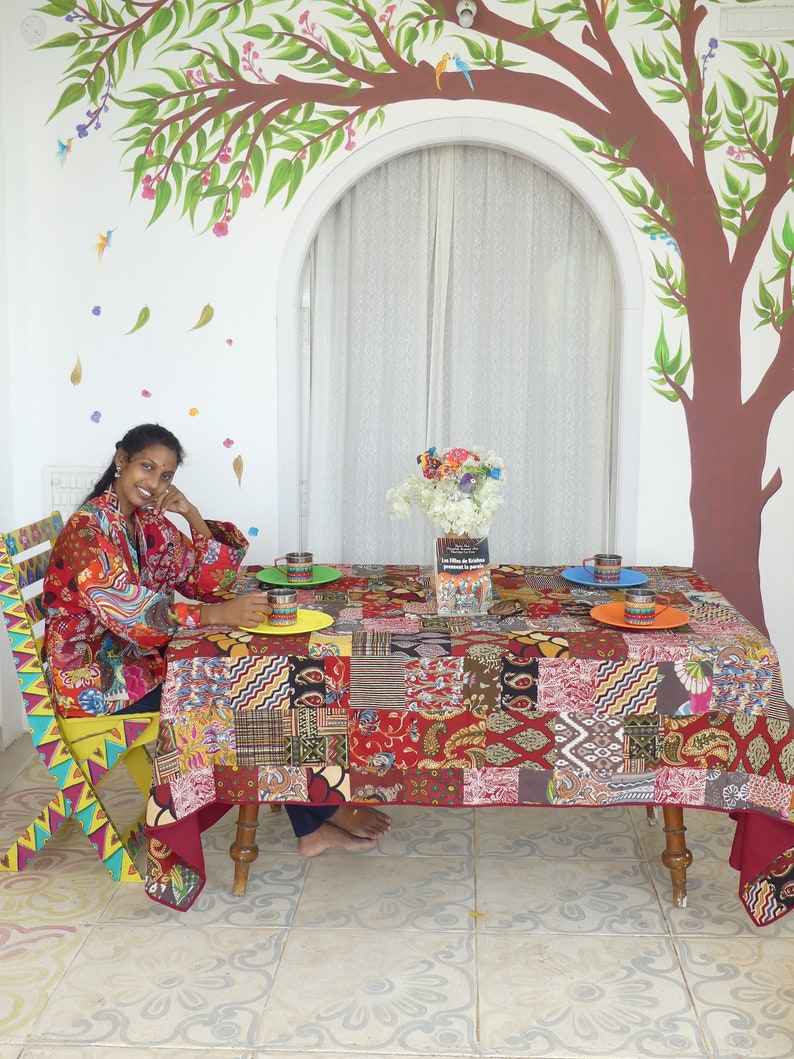 Peut inclure: Une femme est assise &agrave; une table recouverte d'une nappe en patchwork color&eacute;e. La table est dress&eacute;e avec quatre assiettes et tasses color&eacute;es. La femme porte une chemise &agrave; motifs rouges et bleus. L'arri&egrave;re-plan pr&eacute;sente une peinture murale d'un arbre avec des oiseaux.