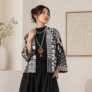 Könnte beinhalten: Schwarz-weiß gemusterte Jacke mit geometrischem Design. Die Jacke wird über einem schwarzen Oberteil und Rock getragen. Eine lange Halskette mit Holzanhänger und eine Perlenkette sind ebenfalls sichtbar.