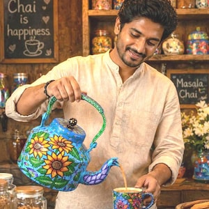May include: A person pouring tea from a blue teapot with sunflower designs into a matching mug. The teapot and mug are decorated with yellow, orange, and green floral patterns. The background includes jars of spices and a chalkboard sign that says "Chai is happiness."