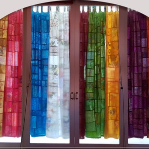 May include: A set of eight sheer curtains in various colors, including orange, red, blue, white, green, yellow, purple, and orange. The curtains are made of a patchwork of fabric squares and are hung on a window.