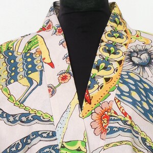 K&ouml;nnte beinhalten: Ein hellfarbener Bademantel mit einem lebendigen, farbenfrohen Muster. Das Design zeigt stilisierte V&ouml;gel, Blumen und Bl&auml;tter in Blau-, Gelb-, Rot- und Gr&uuml;nt&ouml;nen. Der Bademantel hat einen Schalkragen und eine lockere Passform.