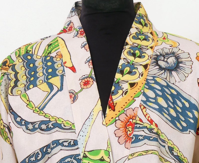 K&ouml;nnte beinhalten: Ein hellfarbener Bademantel mit einem lebendigen, farbenfrohen Muster. Das Design zeigt stilisierte V&ouml;gel, Blumen und Bl&auml;tter in Blau-, Gelb-, Rot- und Gr&uuml;nt&ouml;nen. Der Bademantel hat einen Schalkragen und eine lockere Passform.