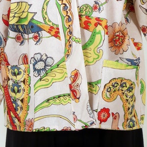 K&ouml;nnte beinhalten: Cremefarbene Jacke mit einem lebendigen Blumen- und Vogelmuster in Rot-, Gelb-, Gr&uuml;n-, Blau- und Oranget&ouml;nen. Die Jacke hat zwei Vordertaschen und lange &Auml;rmel. Der untere Teil der Jacke ist schwarz.