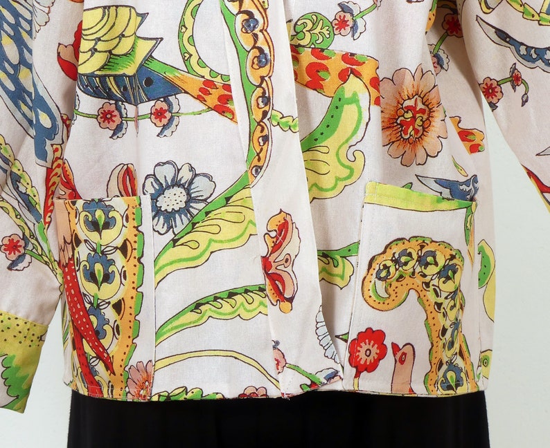 K&ouml;nnte beinhalten: Cremefarbene Jacke mit einem lebendigen Blumen- und Vogelmuster in Rot-, Gelb-, Gr&uuml;n-, Blau- und Oranget&ouml;nen. Die Jacke hat zwei Vordertaschen und lange &Auml;rmel. Der untere Teil der Jacke ist schwarz.