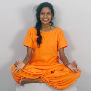 May include: A woman wearing a bright orange outfit sits in a meditative pose with her legs crossed and hands resting on her knees. She is smiling and has long dark hair.