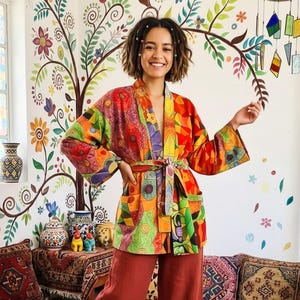 Peut inclure: Une robe de style kimono aux motifs colorés avec une ceinture assortie. La robe présente un mélange vibrant de formes géométriques orange, rouge, vert et violet. Le modèle porte un pantalon couleur rouille. L'arrière-plan comprend une fresque florale et des objets décoratifs.