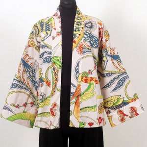 K&ouml;nnte beinhalten: Eine cremefarbene Jacke im Kimono-Stil mit einem lebendigen, farbenfrohen Muster. Das Design zeigt V&ouml;gel, Blumen und abstrakte Formen in Blau-, Gr&uuml;n-, Gelb- und Rott&ouml;nen. Die Jacke hat weite &Auml;rmel und eine lockere Passform.
