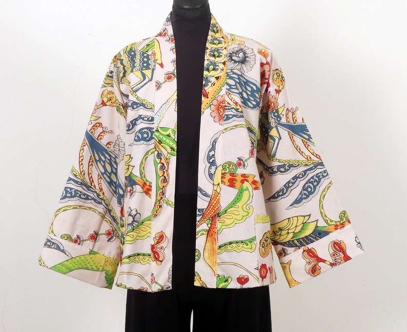 K&ouml;nnte beinhalten: Eine cremefarbene Jacke im Kimono-Stil mit einem lebendigen, farbenfrohen Muster. Das Design zeigt V&ouml;gel, Blumen und abstrakte Formen in Blau-, Gr&uuml;n-, Gelb- und Rott&ouml;nen. Die Jacke hat weite &Auml;rmel und eine lockere Passform.