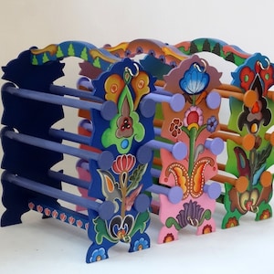 Könnte beinhalten: Drei dekorative Holzregale mit bemalten Blumendesigns. Die Regale sind in Blautönen, Rosa und Grün lackiert. Jedes Regal hat ein einzigartiges Blumendesign und ist mit kleinen runden Knöpfen verziert.